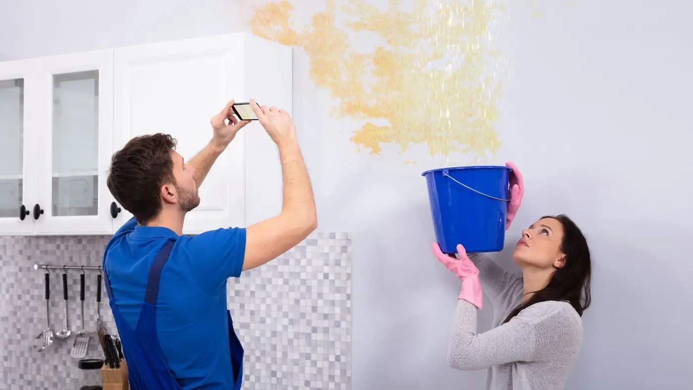 Homeowner photographing a ceiling leak while another person holds a bucket